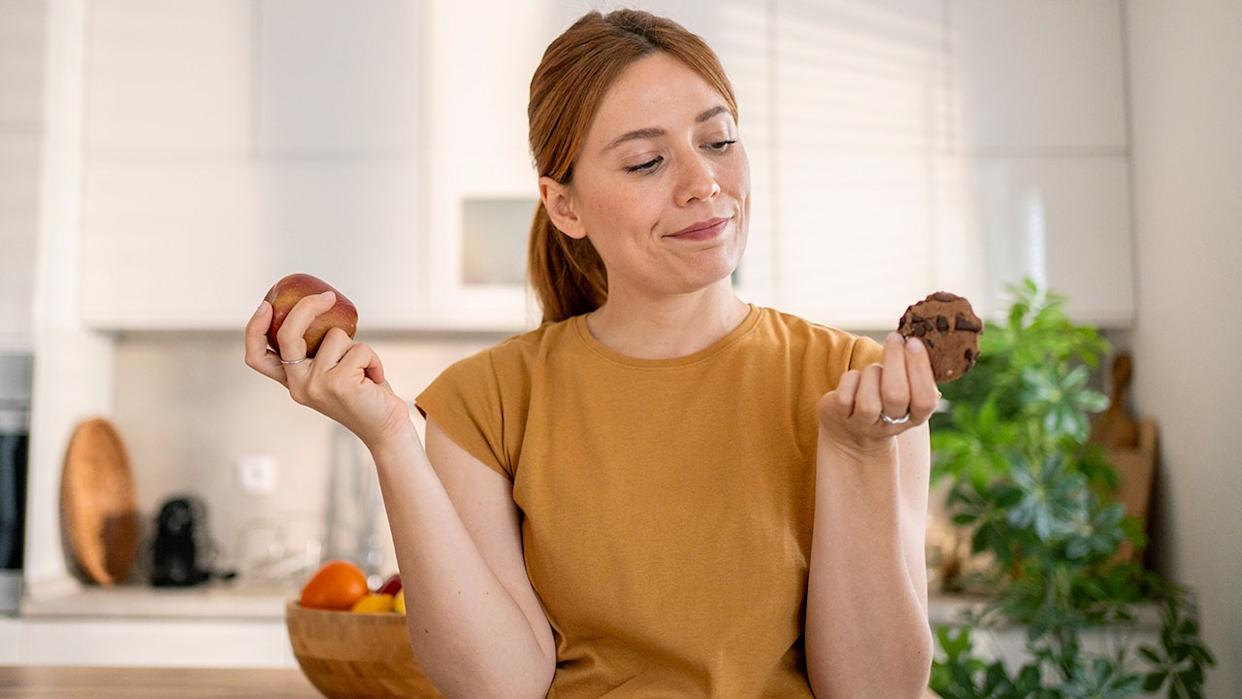 Young woman deciding between eating a red apple or a chocolate cookie, holding them in her hands, sitting in a modern kitchen, struggling with a healthy lifestyle.