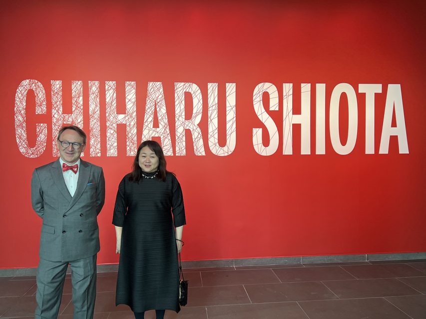 Two people are standing in front of a red wall with large white letters written on it. "CHIHARU SHIOTA.