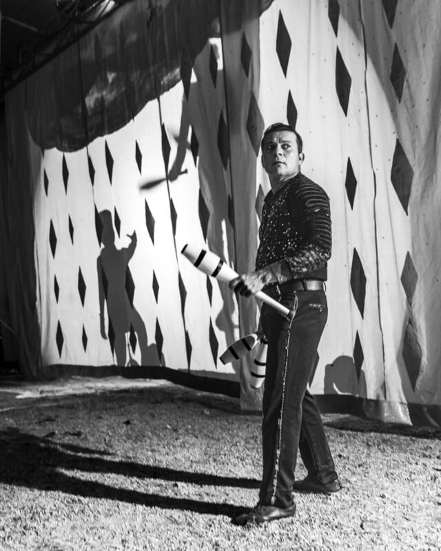 A man in a sequined shirt juggles clubs in front of a diamond-patterned curtain, casting dramatic shadows against a black-and-white circus backdrop.
