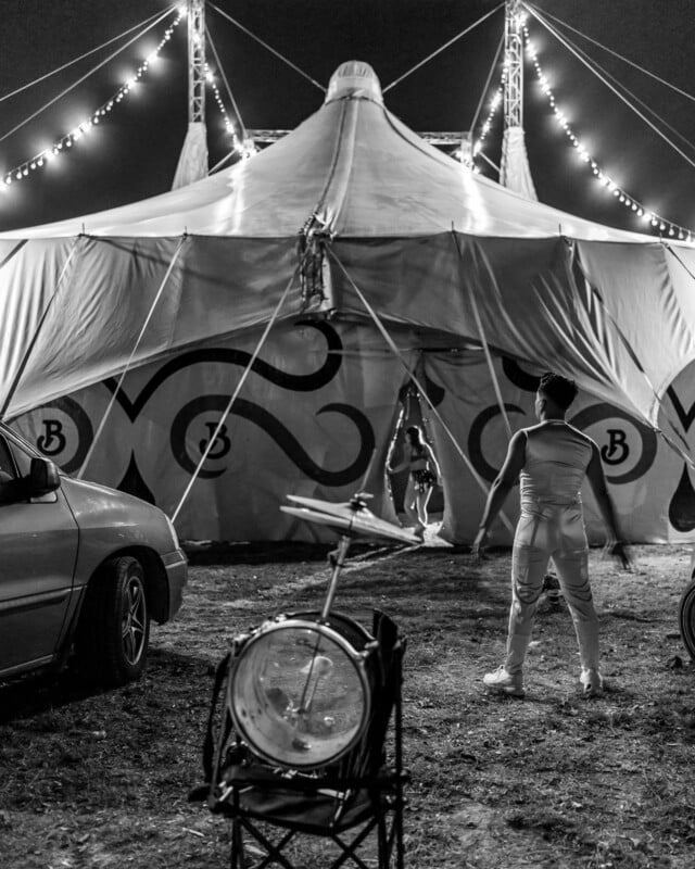 Black and white photo of a circus tent at night. A costumed performer stands in front, facing off against an acrobat hanging from a rope at the entrance. Oil drums and parked cars can be seen in the foreground.