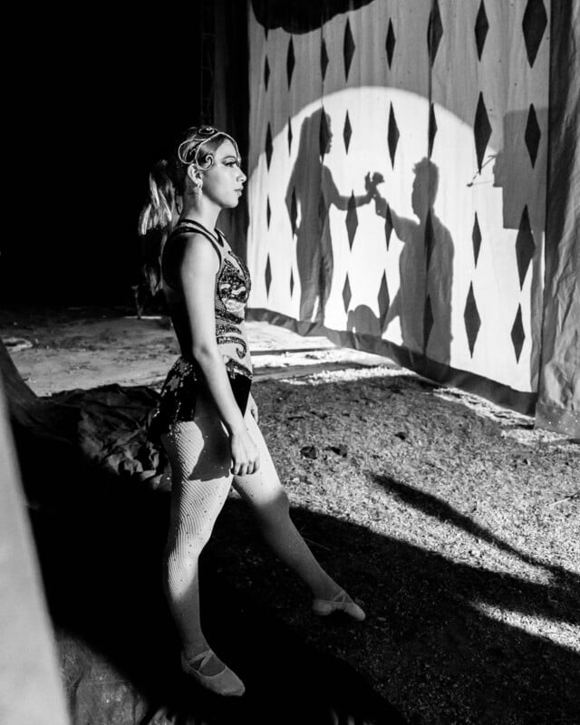 A costumed circus performer stands backstage, staring out at a stage shadowed by diamond-patterned curtains and spotlights. The scene is black and white.