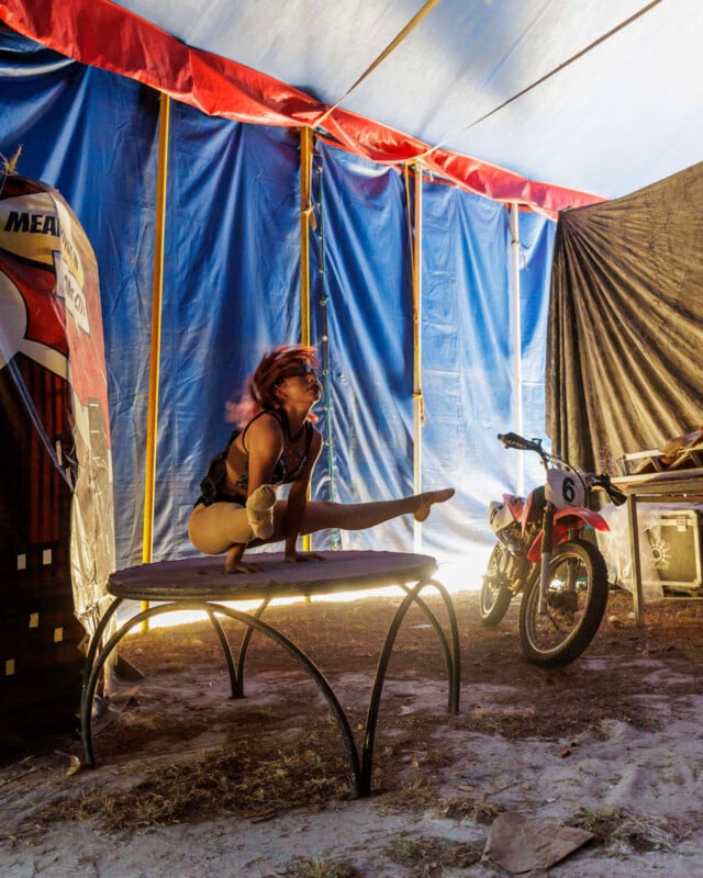 Acrobats perform at a round table inside a circus tent, with motorcycles and equipment in the background. A blue and red cloth wall is visible, and sunlight is shining in from the left side.