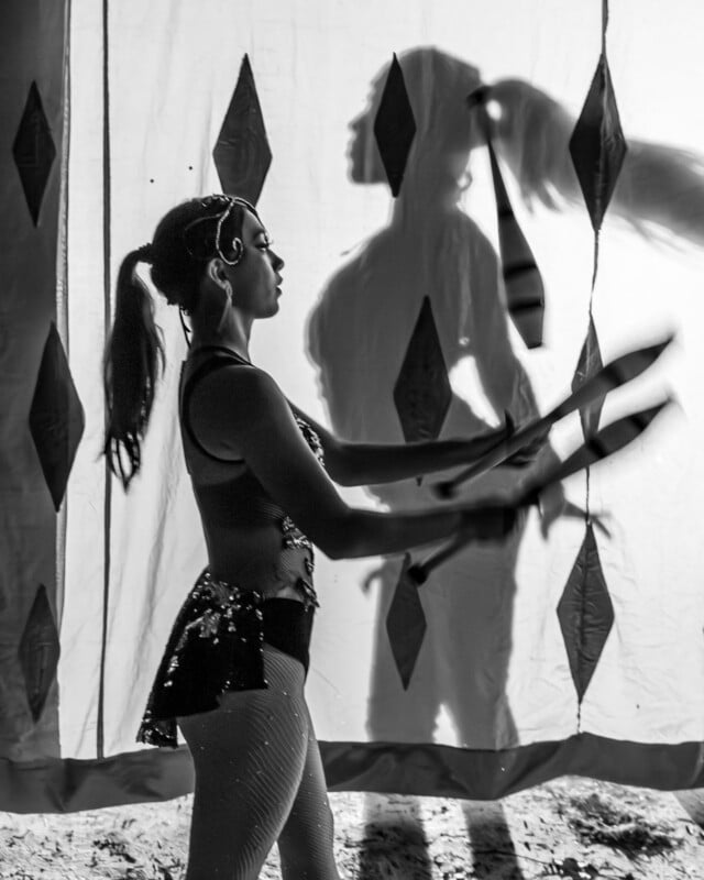 A costumed woman juggles clubs in front of patterned curtains, casting a large shadow behind her. The scene is black and white.