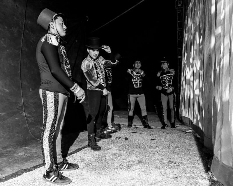 Five male circus performers wearing decorative costumes and top hats stand under a tent backstage. Some people adjust their costumes, while others wait with dramatic lighting and shadows cast on the walls of their tents.