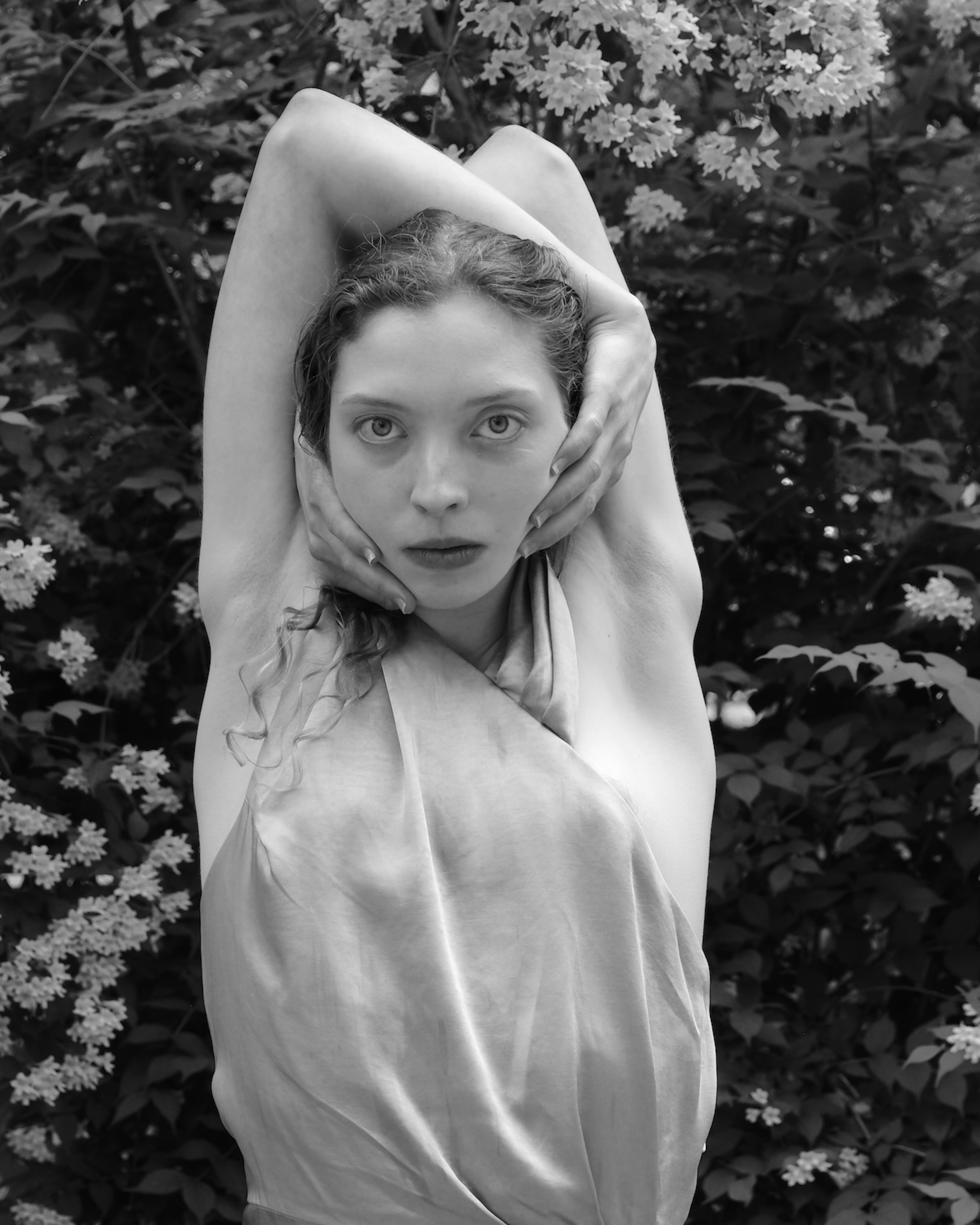 In this black and white portrait taken among the shrubs of a flowering garden, a young woman looks directly into the camera with her arms raised above her head.