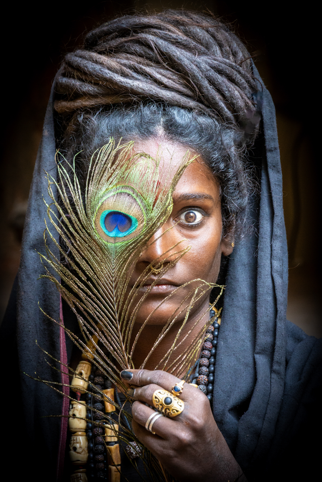 A figure wrapped in a gray cloth head holds a bright peacock feather over half its face, layered with a beaded necklace and ornate ring, revealing one intense eye.