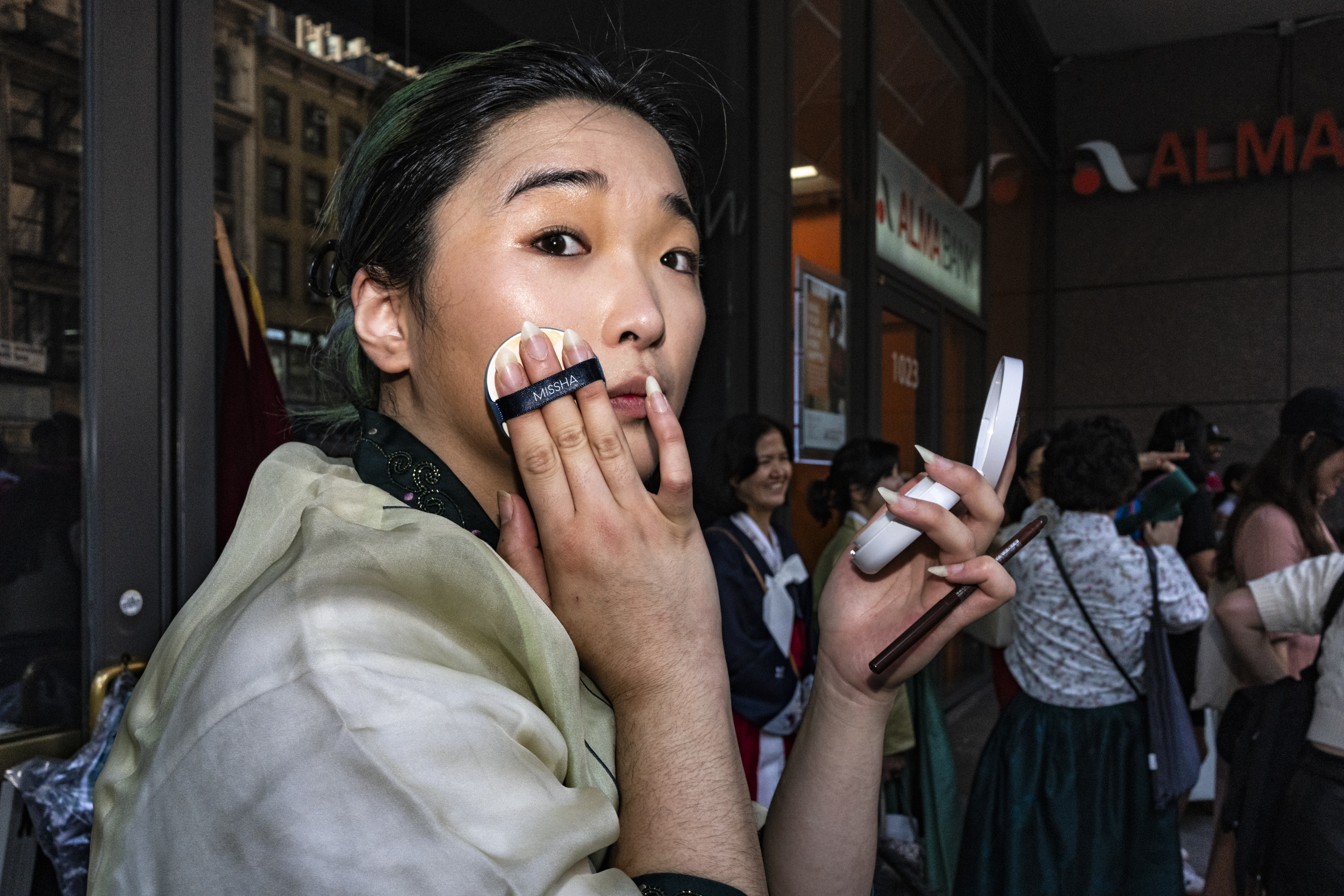 A young man with dark greenish hair is standing on a busy street in the city, holding a small mirror and applying foundation with a Missha cushion compact.
