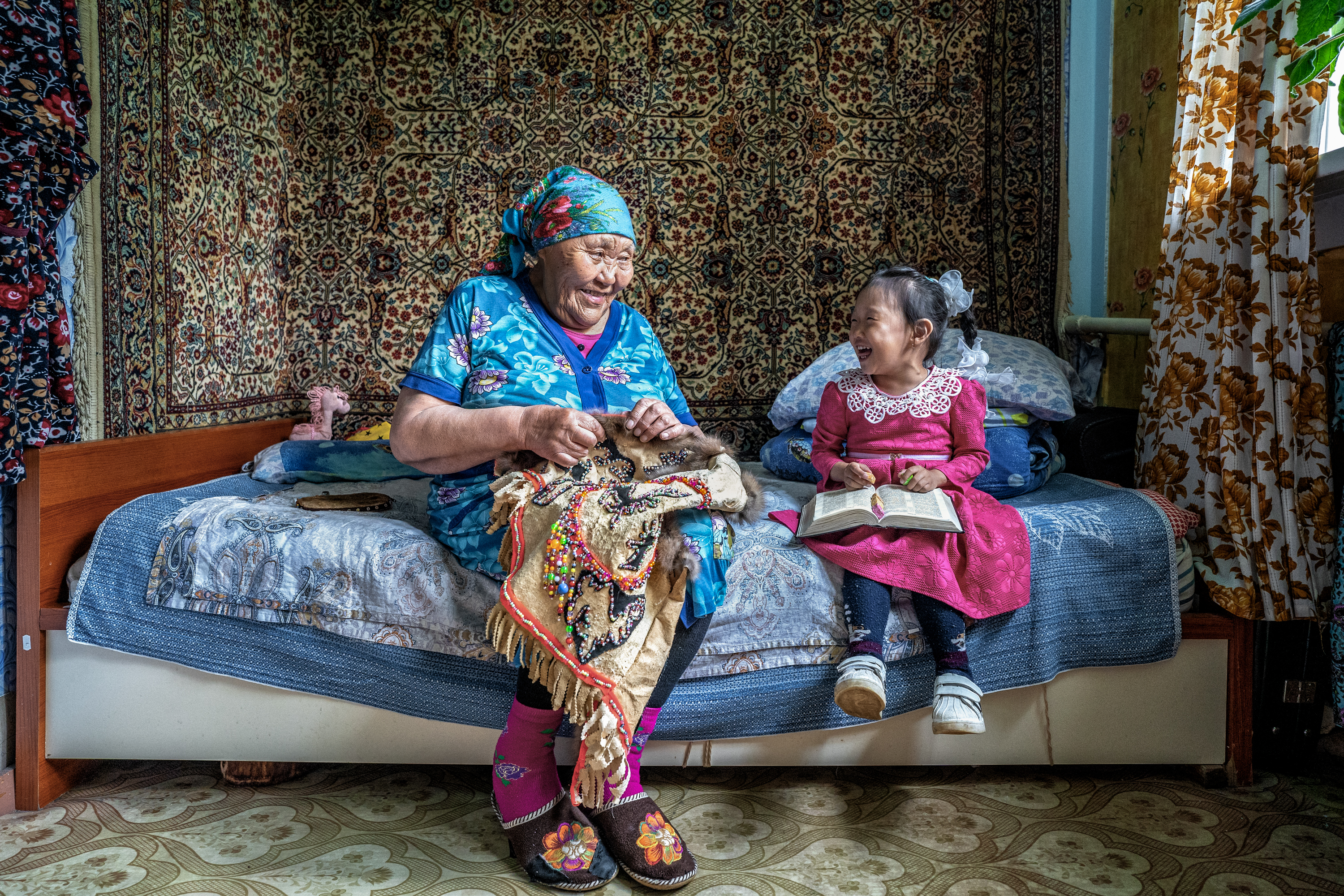 An elderly woman sitting on her bed sewing richly decorated textiles smiles at a young girl sitting next to her with an open book. The two share a warm moment in a cozy patterned room.