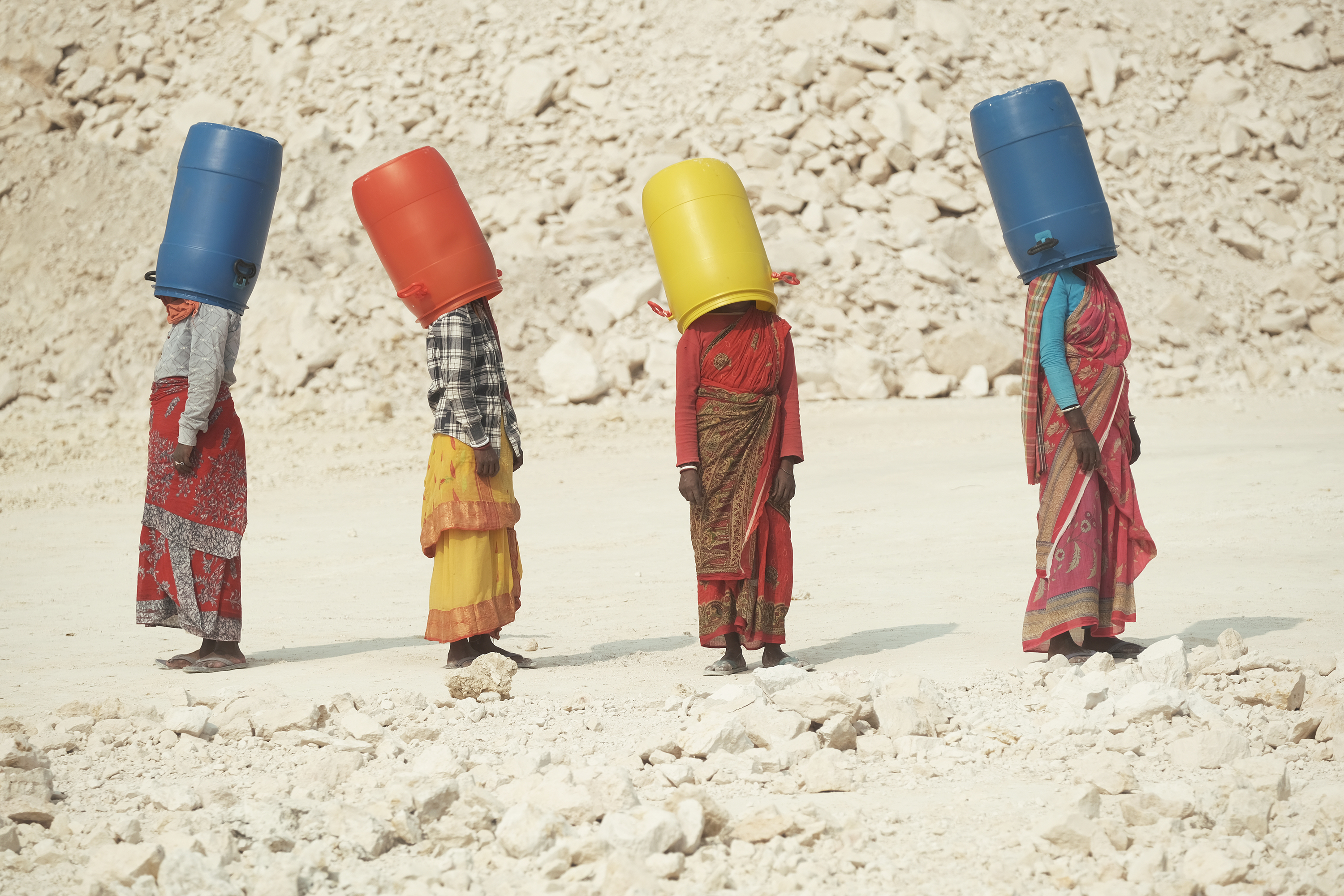 Four women in colorful saris stand in a line against a pale, rocky landscape, each balancing on a large plastic barrel. Blue, orange, yellow, blue - Cover your head to hide your face.