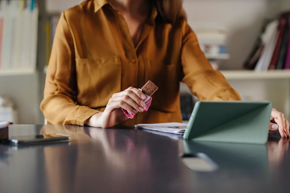 Anonymous businesswoman eating protein bar while sitting in office and video calling with colleagues