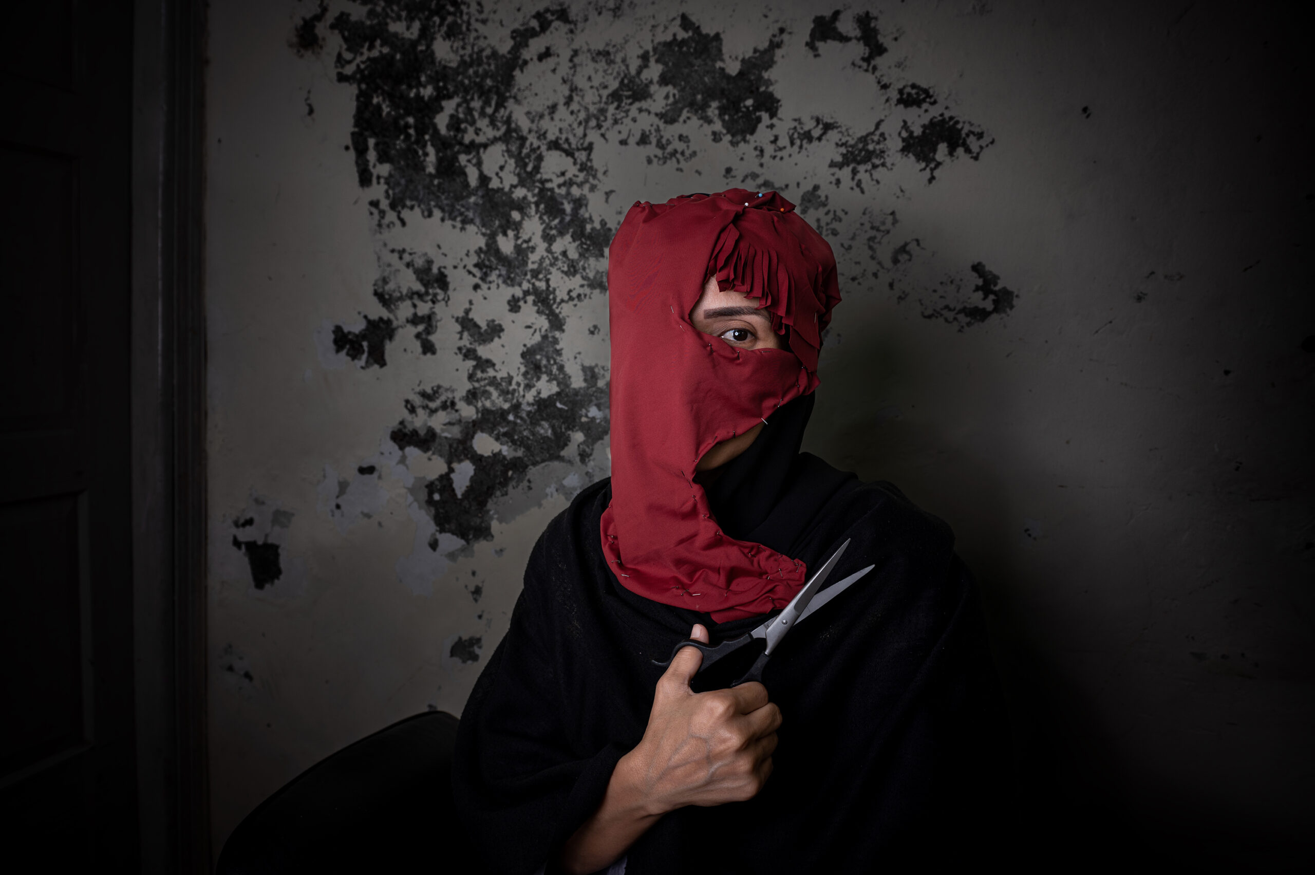 A figure, whose face is almost completely covered by a torn red scarf, holds a pair of scissors to his chest and stares with one eye at a dark, peeling wall.