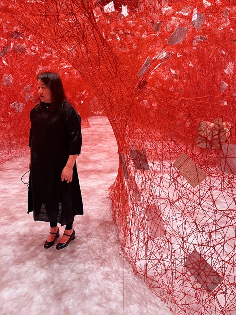 A woman stands near an intricate installation of red thread and hanging paper woven between tree-like structures in the gallery space.