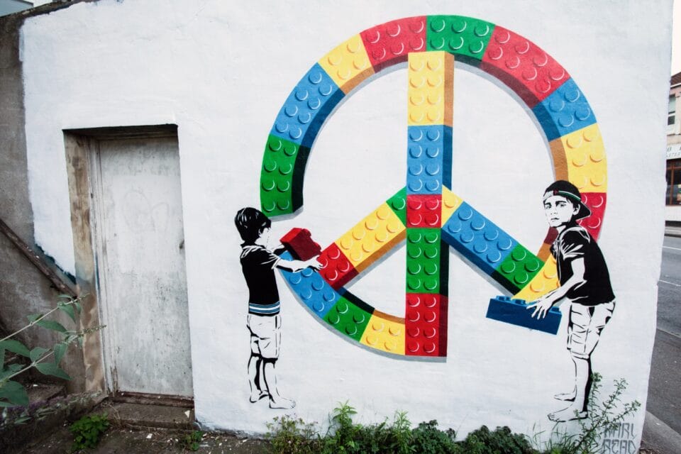 A street art mural of a peace sign made of colorful Lego blocks and two children assembling it.