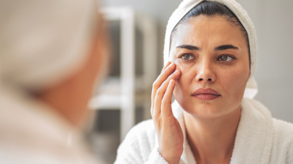 A woman wearing a white robe and hair towel touches the skin around her eyes and stares at her reflection in the mirror with a worried expression.